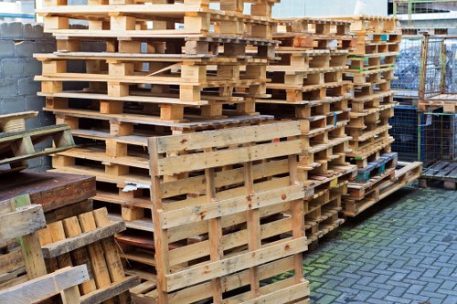 A large stack of untreated wooden pallets arranged in several columns and rows, with some pallets leaning slightly to the side. The pallets are located outdoors on a paved area, with a background that includes a grey brick wall and a metal fence. The pallets vary in size and appear to be used for transporting goods, featuring horizontal slats supported by vertical blocks. The scene is illuminated by natural daylight, highlighting the rough texture and light brown colour of the wood. To the left, there are a few more pallets and wooden crates, some showing signs of weathering. This scene is typical of a rubbish removal or waste management context, as managed by Garden Clearance Hackney, serving the local area and facilitating the proper disposal of waste materials such as used pallets.