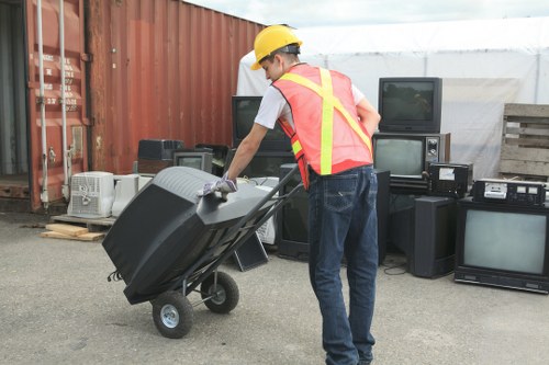 A person wearing a yellow safety helmet and a high-visibility vest is pushing a black wheelbarrow filled with electronic waste, including monitors and small appliances. The individual is dressed in a white T-shirt, blue jeans, and work gloves. The scene is set outdoors on a paved area, with a red corrugated metal fence on the left and a stack of various discarded electronic devices, such as televisions and audio equipment, arranged against the fence. In the background, there is a white structure covered with a tarp, suggesting a waste collection or recycling site. The overall environment indicates a professional rubbish removal operation, likely associated with Garden Clearance Hackney, focusing on electronics disposal as part of garden, house, or commercial clearance services in London or East London boroughs.