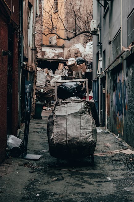 A street scene showing a white waste collection truck with an open rear compartment clearly designed for rubbish collection, positioned on a cobblestone road next to a row of old, weathered buildings