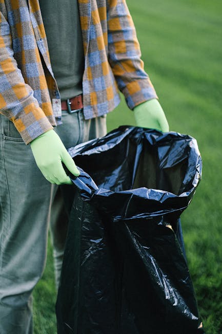 A pile of black rubbish bags and discarded packaging materials stacked haphazardly in front of a red metal door set into a textured, off-white exterior wall. The bags are made of plastic and appear fu