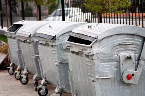 Four large, grey metal wheelie bins with closed lids are positioned in a row on a paved surface, likely outside a property in Hackney. The bins are made of galvanized steel with ribbed sides and rounded top edges, and each has four small black wheels at the base for mobility. The lids are slightly curved, and one bin's lid is partially open, revealing a dark interior. The background features a black metal fence with vertical bars, and beyond that, a street scene with parked white vehicles and green trees, indicating an urban or residential environment. The lighting suggests daytime with natural light illuminating the scene, typical of a typical rubbish collection area used by Garden Clearance Hackney for waste removal services. The setup aligns with typical rubbish management practices in the Hackney area, reflecting the company's focus on waste collection and clearance services.