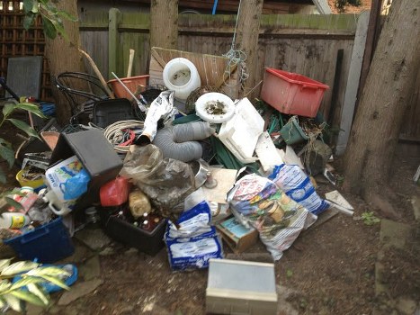 A large pile of assorted household rubbish and garden waste is collected on a patch of earth near a wooden fence in an outdoor area. The heap includes plastic buckets, bags of trash, cardboard boxes, plastic containers, and various miscellaneous waste items. Some objects are stacked or leaning against each other, with parts of the rubbish spilling onto the ground. In the background, there are wooden fence panels and tree trunks, indicating an outdoor setting, possibly a garden or backyard in Hackney. The items appear to be awaiting disposal or removal, characteristic of a garden clearance service, with a mix of durable plastics and softer materials. The scene is lit by natural daylight, providing clear visibility of the clutter and textures, emphasizing the need for professional rubbish removal such as that provided by Garden Clearance Hackney.