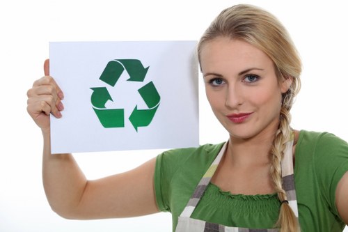 A woman with blonde hair styled in a side braid, wearing a green top and a striped apron, holds up a white sheet of paper displaying a green recycling symbol composed of three arrows forming a triangle, with her right arm extended forward. She stands against a plain white background, capturing a clear and neutral expression, directly facing the camera. The focus is on her and the recycling symbol, and her attire suggests involvement in eco-friendly or waste management initiatives, relevant to rubbish removal services in London areas such as Hackney. The image emphasizes environmental awareness and sustainability, aligning with professional waste disposal services provided by companies like Garden Clearance Hackney. The setting appears studio-lit, ensuring the colors are vibrant and the details sharp, with no visible clutter or background distractions, making it suitable for a website focused on rubbish and garden clearance solutions within the Hackney district.